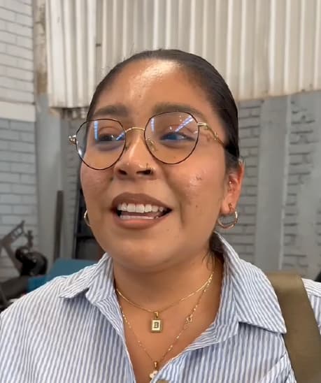 Una persona con gafas, con camisa de rayas y collares dorados, y sonriendo en un interior con una pared de metal corrugado de fondo.